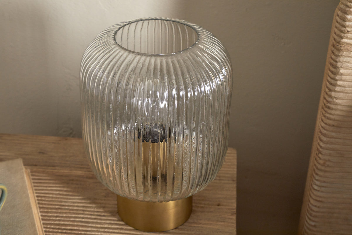 The Rizal Fluted Glass Bedside Table Lamp by nkuku, featuring a cylindrical clear glass design and antique brass base, rests on a wooden surface with sunlight casting soft shadows beside a beige wall and woven chair.