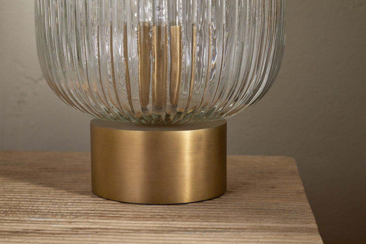 The nkuku Rizal Fluted Glass Bedside Table Lamp in Antique Brass & Clear features a ribbed, clear base and provides ambient lighting on a light wooden surface against a plain beige wall.