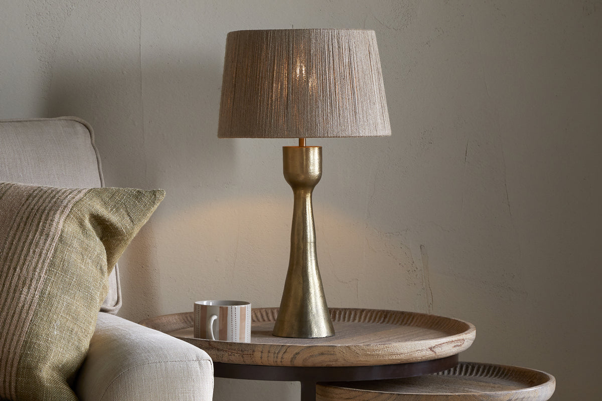 A beige armchair with a textured cushion sits by a round wooden side table. On the table are a ceramic mug and the nkuku Marpha Antique Brass Table Lamp, featuring a beige fabric shade that casts warm accent lighting on the neutral wall.