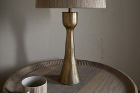 The nkuku Marpha Antique Brass Table Lamp with a beige fabric shade sits on a round wooden tray table, casting accent light. To the left is a white mug with beige and gray stripes, set against a textured off-white wall.