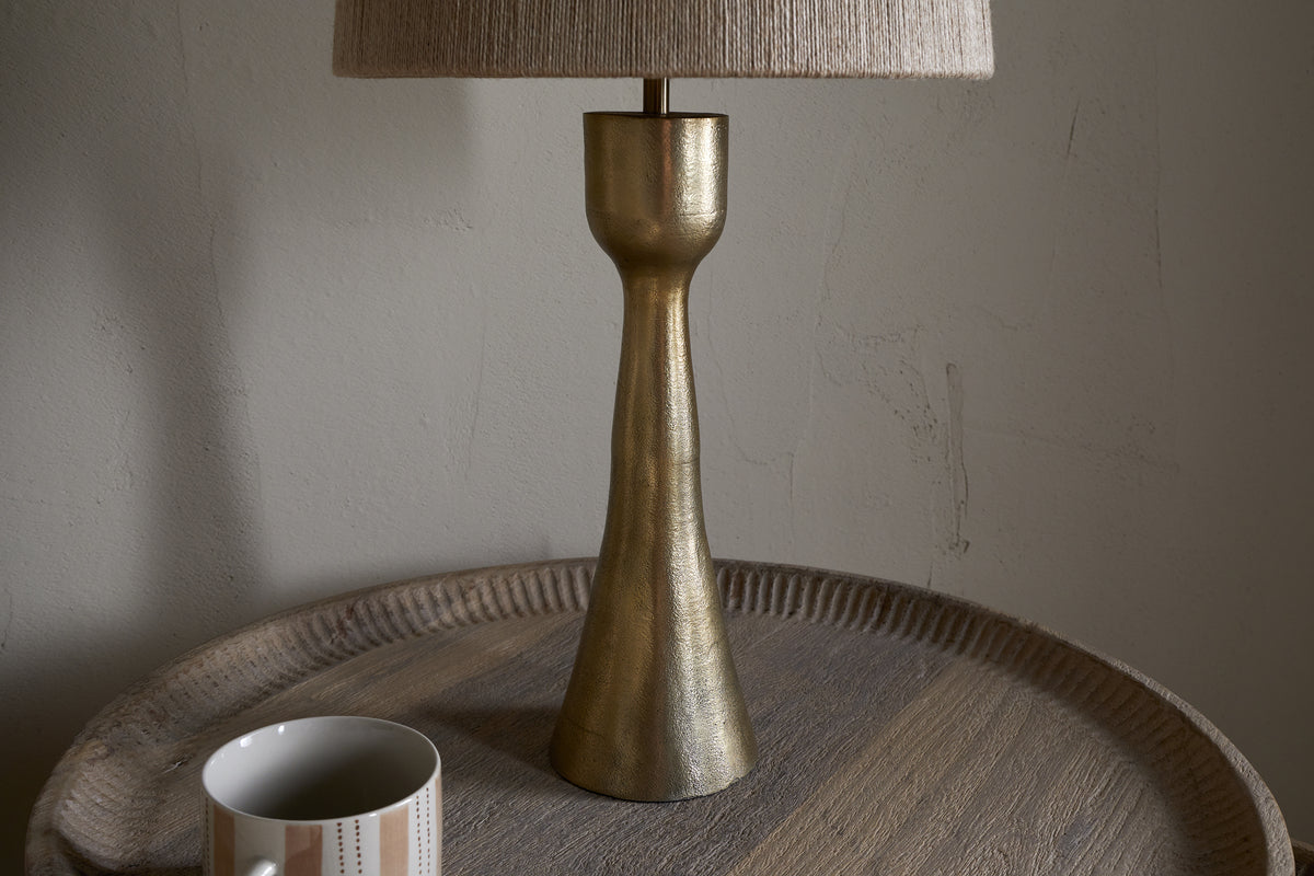 The nkuku Marpha Antique Brass Table Lamp with a beige fabric shade sits on a round wooden tray table, casting accent light. To the left is a white mug with beige and gray stripes, set against a textured off-white wall.