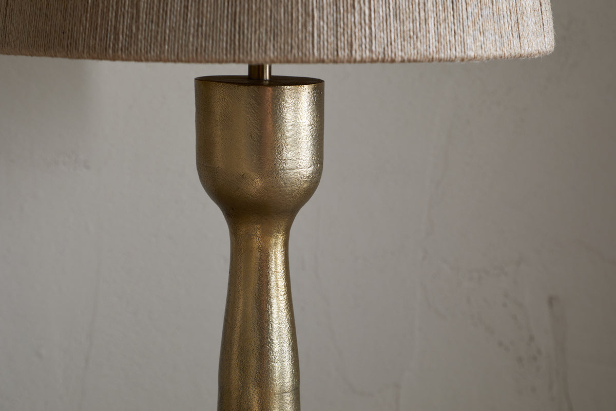A close-up of the nkuku Marpha Antique Brass Table Lamp shows its textured gold base and beige fabric shade. Ideal as accent lighting, it stands against a plain, light wall with subtle marks. Only the upper base and lower edge of the shade are visible.