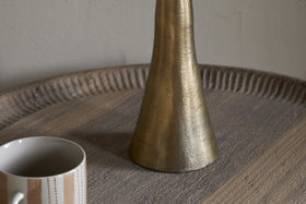 The Marpha Antique Brass Table Lamp by nkuku, featuring a textured metallic gold cone shape, rests on a round ridged wooden tray. To the left is a white ceramic mug with beige stripes and dots, set against a plain light wall.