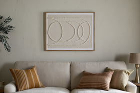 A beige sofa with three earth-toned pillows sits against a light wall, topped by the nkuku Mauri Frayed Wall Art—neutral-textured abstract circles in a wooden frame. A lamp and plant are partially visible on either side.