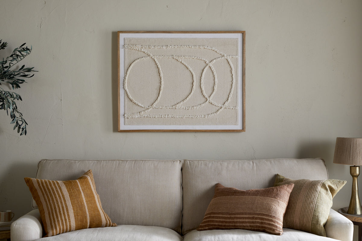 A beige sofa with three earth-toned pillows sits against a light wall, topped by the nkuku Mauri Frayed Wall Art—neutral-textured abstract circles in a wooden frame. A lamp and plant are partially visible on either side.