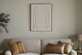 A beige sofa with two striped cushions sits against a light wall. Above hangs the Mauri Frayed Wall Art - Neutral by nkuku, its textured, curved lines framed. Side tables with a lamp and plant complete the cozy, neutral-toned decor.