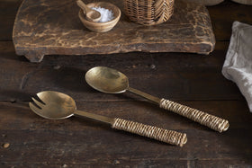 The Abani Salad Servers by nkuku—antique brass with woven rattan handles—rest on a rustic wood table, joined by a cutting board, seagrass basket, salt bowl, and cloth napkin.
