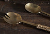 The nkuku Abani Salad Servers in antique brass feature a spoon and two-tined fork-spoon with square handles. Handcrafted with seagrass twine on one handle, their slightly tarnished surfaces add rustic charm.
