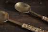 The Abani Salad Servers by nkuku feature antique brass and square handles wrapped in natural twine, their matte finish showcasing handcrafted charm as they rest on rustic, weathered wood.
