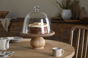 A round cake with cream and chocolate topping sits on the nkuku Vivan Glass Cake Stand & Dome - Natural, atop a rustic wooden table set with serveware, including a striped cup, saucer, and teapot. A wooden chair and decor appear in the background.