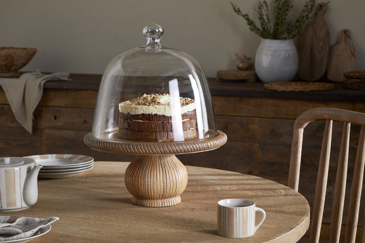 A round cake with cream and chocolate topping sits on the nkuku Vivan Glass Cake Stand & Dome - Natural, atop a rustic wooden table set with serveware, including a striped cup, saucer, and teapot. A wooden chair and decor appear in the background.