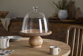 The nkuku Vivan Glass Cake Stand & Dome - Natural sits on a round table, paired with a striped mug, matching plate and teapot, and cloth napkin. Rustic decor and ceramics accent the wooden sideboard behind.
