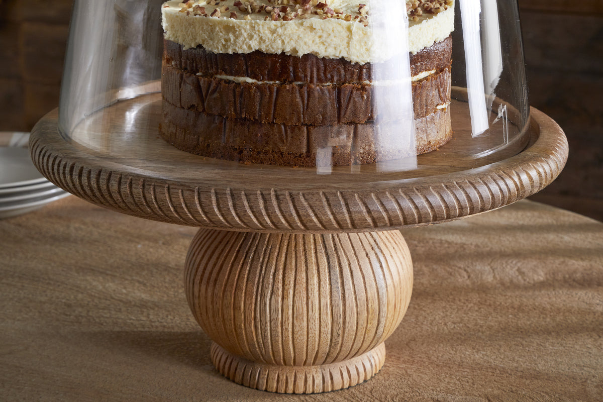 A layered cake with white frosting and nuts sits on the nkuku Vivan Glass Cake Stand & Dome - Natural, featuring a carved mango wood base and clear glass cover, with serveware stacked in the blurred background.