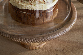 A partially visible layered cake with cream frosting and nut crumbles sits on the nkuku Vivan Glass Cake Stand & Dome - Natural, featuring a grooved mango wood base and glass cover, placed on a wooden table.