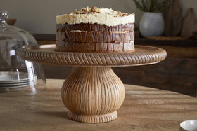 A chocolate layer cake with thick white frosting and chopped nuts sits on the nkuku Vivan Glass Cake Stand & Dome - Natural, displayed on a round wooden table with rustic pottery and plates visible in the softly blurred background.