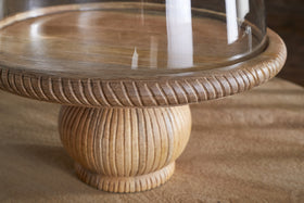 The Vivan Glass Cake Stand & Dome – Natural by nkuku features a mango wood base with carved texture and fine grain, topped with a clear dome, showcasing the light finish against a softly blurred background.