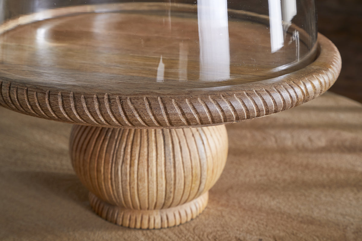 The Vivan Glass Cake Stand & Dome – Natural by nkuku features a mango wood base with carved texture and fine grain, topped with a clear dome, showcasing the light finish against a softly blurred background.