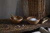 The nkuku Daruka Salt & Pepper Dipping Bowls with Spoons - Natural rest on a rustic wooden surface, one cradling coarse sea salt, the other empty but for a spoon. Scattered salt crystals and cozy table accents enhance the inviting scene.