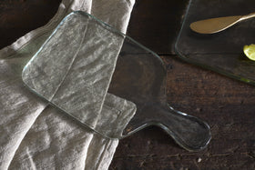 The nkuku Komal Serving Platter - Clear with a handle sits on a beige linen atop dark wood, while another stylish platter with a gold butter knife and lime slice appears in the top right corner.