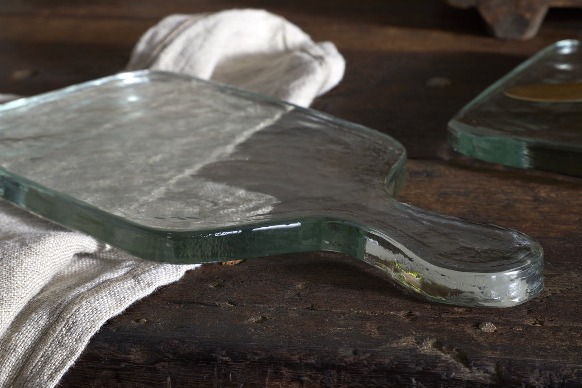 The Komal Serving Platter - Clear by nkuku features thick, clear glass with smooth edges and a handle. Resting on a rustic wooden surface beside a beige cloth, its shiny finish adds style to any serveware collection.