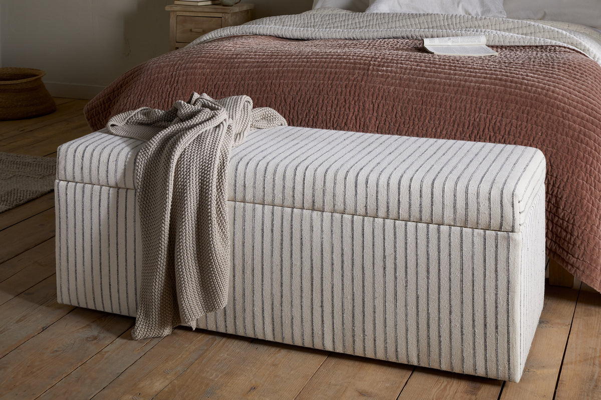 The nkuku Sanja Stripe Cotton & Linen Upholstered Storage Bench in Grey sits at the foot of a bed with a textured reddish-brown quilt and white sheets, adding practical bedroom storage to the cozy, minimalist space with neutral decor and wooden floors.