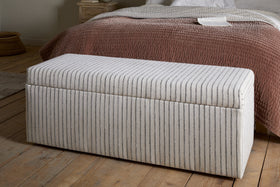 The nkuku Sanja Stripe Cotton & Linen Upholstered Storage Bench - Grey, featuring vertical beige and grey stripes, adds stylish storage at the foot of the bed with mauve-brown quilt and white bedding in a cozy bedroom.