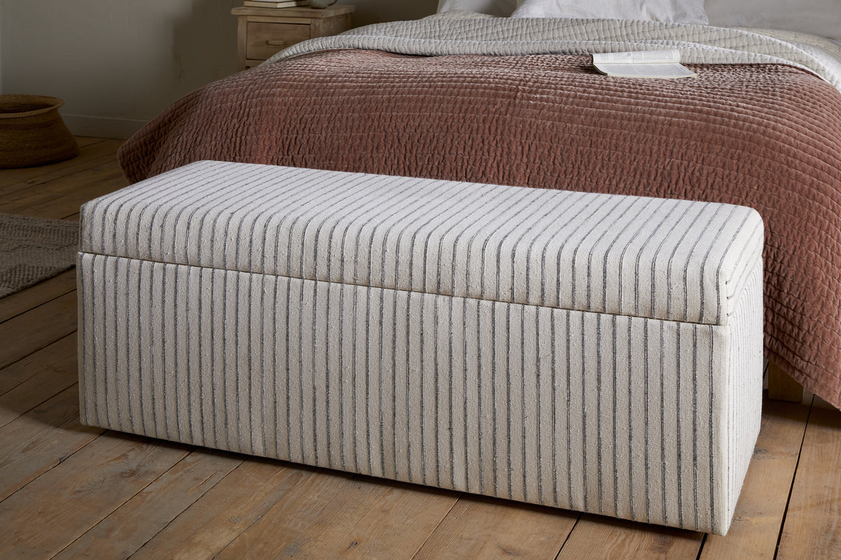 The nkuku Sanja Stripe Cotton & Linen Upholstered Storage Bench - Grey, featuring vertical beige and grey stripes, adds stylish storage at the foot of the bed with mauve-brown quilt and white bedding in a cozy bedroom.