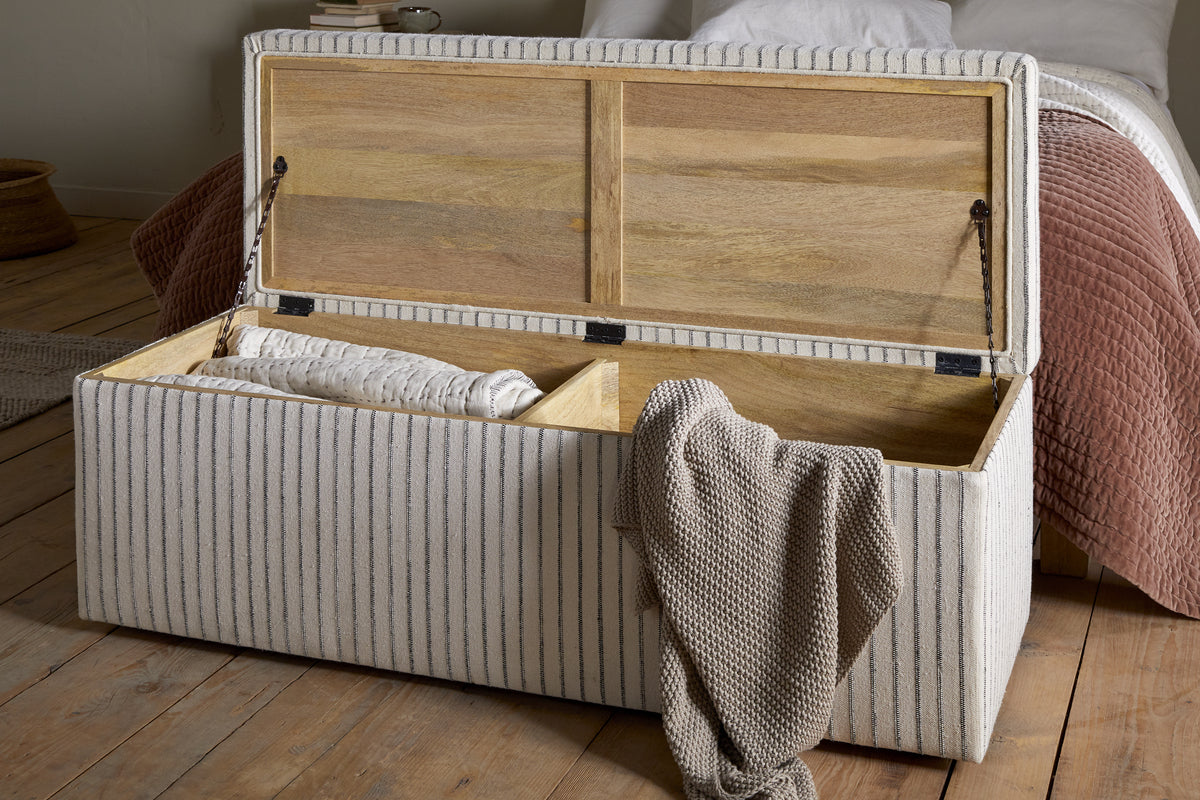 The nkuku Sanja Stripe Cotton & Linen Upholstered Storage Bench - Grey sits on a wooden floor at the foot of a bed with white and taupe bedding, revealing handy bedroom storage for blankets and throws inside.