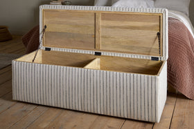 The nkuku Sanja Stripe Cotton & Linen Upholstered Storage Bench in grey features a hinged lid, two wooden compartments, and light fabric with thin gray stripes, offering stylish bedroom storage beside a bed.