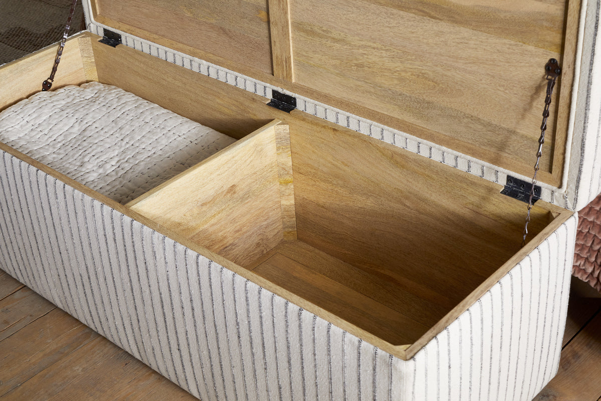 The nkuku Sanja Stripe Cotton & Linen Upholstered Storage Bench in Grey features a light wood interior with two sections—one storing a white quilted blanket, the other empty. Its striped fabric and sturdy metal hinges rest elegantly on a wooden floor.