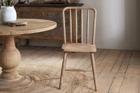 The Om Mango Wood Dining Chair - Natural by nkuku features three vertical backrest slats and stands on a light wooden floor beside a round table, with a woven rug beneath and a wooden sideboard against a white wall in the background.