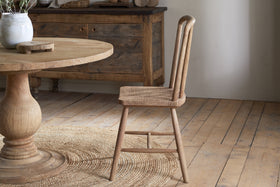 The Om Mango Wood Dining Chair - Natural by nkuku, with its curved backrest, stands by a round wooden table on a woven rug. A rustic floor and decorated sideboard enhance the warm, cozy ambiance.