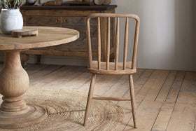 The Om Mango Wood Dining Chair - Natural by nkuku features a slatted back and rustic finish, placed on a round woven rug beside a wooden pedestal table. A wood sideboard and light wood plank floors complete the cozy, farmhouse-inspired setting.