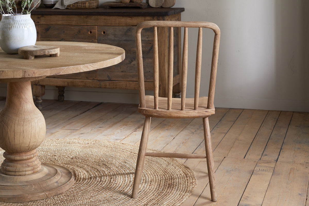 The Om Mango Wood Dining Chair - Natural by nkuku features a slatted back and rustic finish, placed on a round woven rug beside a wooden pedestal table. A wood sideboard and light wood plank floors complete the cozy, farmhouse-inspired setting.