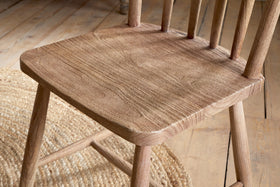 A close-up of the nkuku Om Mango Wood Dining Chair in natural finish shows its textured seat and straight back spindles. The chair stands on a light wood floor next to a round woven jute rug, creating a rustic, airy atmosphere.