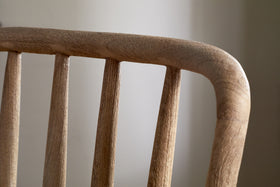 Close-up of the upper backrest of the nkuku Om Mango Wood Dining Chair - Natural, featuring four rounded vertical spindles and a curved top rail in smooth, natural-finish wood against a softly blurred neutral background.