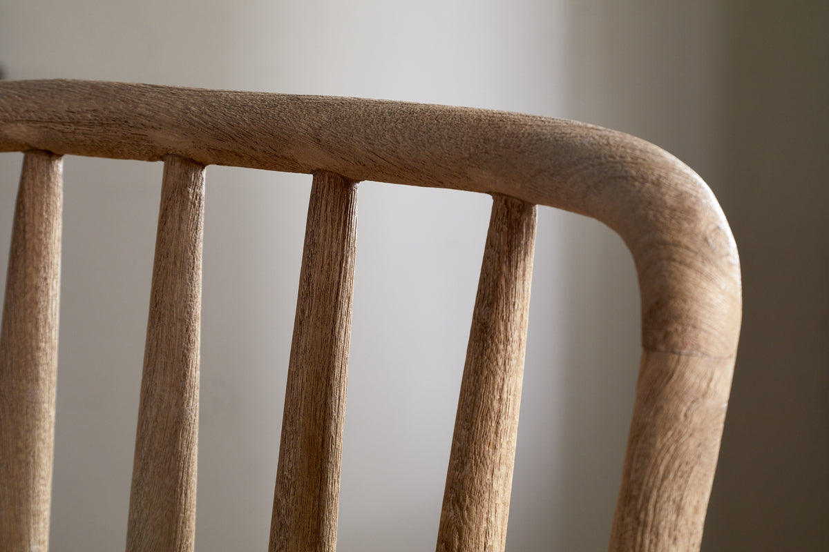 Close-up of the upper backrest of the nkuku Om Mango Wood Dining Chair - Natural, featuring four rounded vertical spindles and a curved top rail in smooth, natural-finish wood against a softly blurred neutral background.