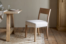 The nkuku Nidhi Linen Upholstered Dining Chair in natural mango wood with off-white striped linen stands by a matching table set on a woven rug atop a rustic wooden floor, with plates, glasses, and a napkin arranged neatly on the tabletop.