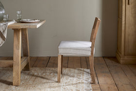 The nkuku Nidhi Linen Upholstered Dining Chair in natural mango wood sits beside a matching table set with plates, glasses, and a striped runner, all arranged on a fringed beige rug over wooden floorboards by a light wall.