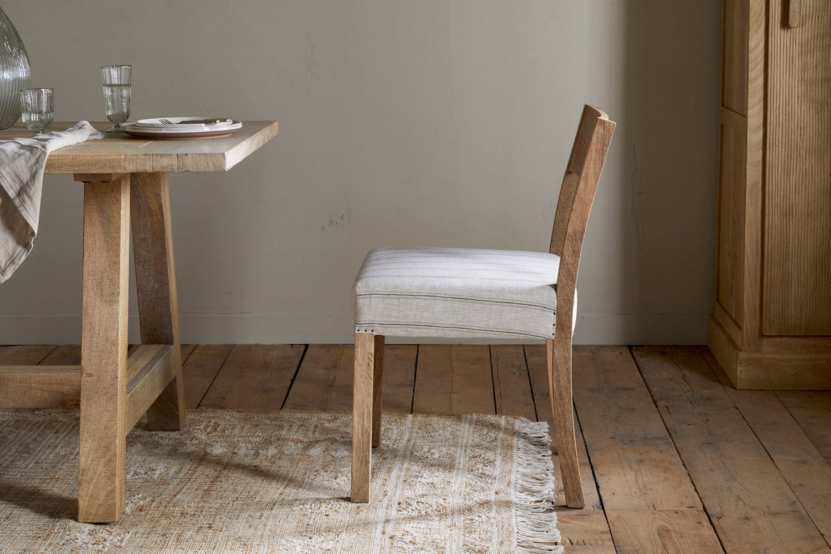 The nkuku Nidhi Linen Upholstered Dining Chair in natural mango wood sits beside a matching table set with plates, glasses, and a striped runner, all arranged on a fringed beige rug over wooden floorboards by a light wall.