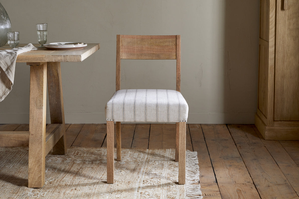 The nkuku Nidhi Linen Upholstered Dining Chair in natural mango wood with a white, subtly striped linen cushion stands by a matching table on a woven rug over wood floors in a neutral room.
