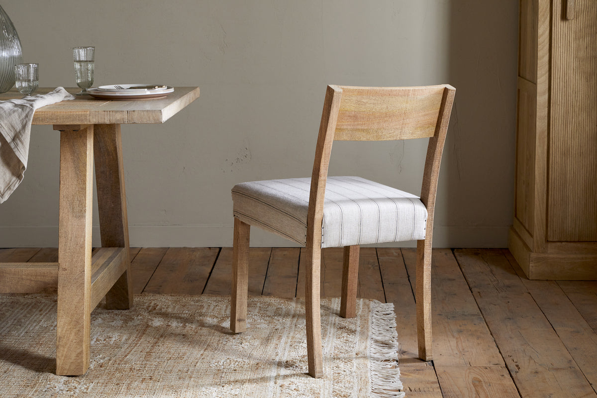 The nkuku Nidhi Linen Upholstered Dining Chair - Natural, with beige striped linen and solid mango wood, stands beside a matching table on a woven rug over rustic flooring, bathed in natural light in a neutral-toned room.