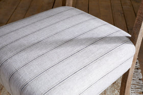 The Nidhi Linen Upholstered Dining Chair - Natural by nkuku features light gray linen with thin dark stripes, solid mango wood legs, and is shown on a wooden floor with part of a patterned rug visible in the corner.