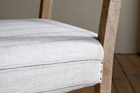 Close-up of the nkuku Nidhi Linen Upholstered Dining Chair—natural mango wood frame, cushioned seat in light beige linen with thin dark stripes, and studded trim. Wooden floorboards and a plain wall set the scene.