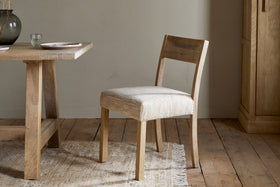 The Zoha Cotton & Jute Upholstered Dining Chair - Natural by nkuku sits beside a matching table set with plates and a glass, on a fringed rug over rustic wood flooring, with a cabinet and neutral wall in the background.