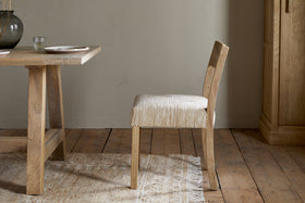 The Zoha Cotton & Jute Upholstered Dining Chair - Natural by nkuku features a textured beige seat and backrest beside a matching wooden dining table, set on a light rug over wooden floors, with a cupboard and ceramic vase in the background.