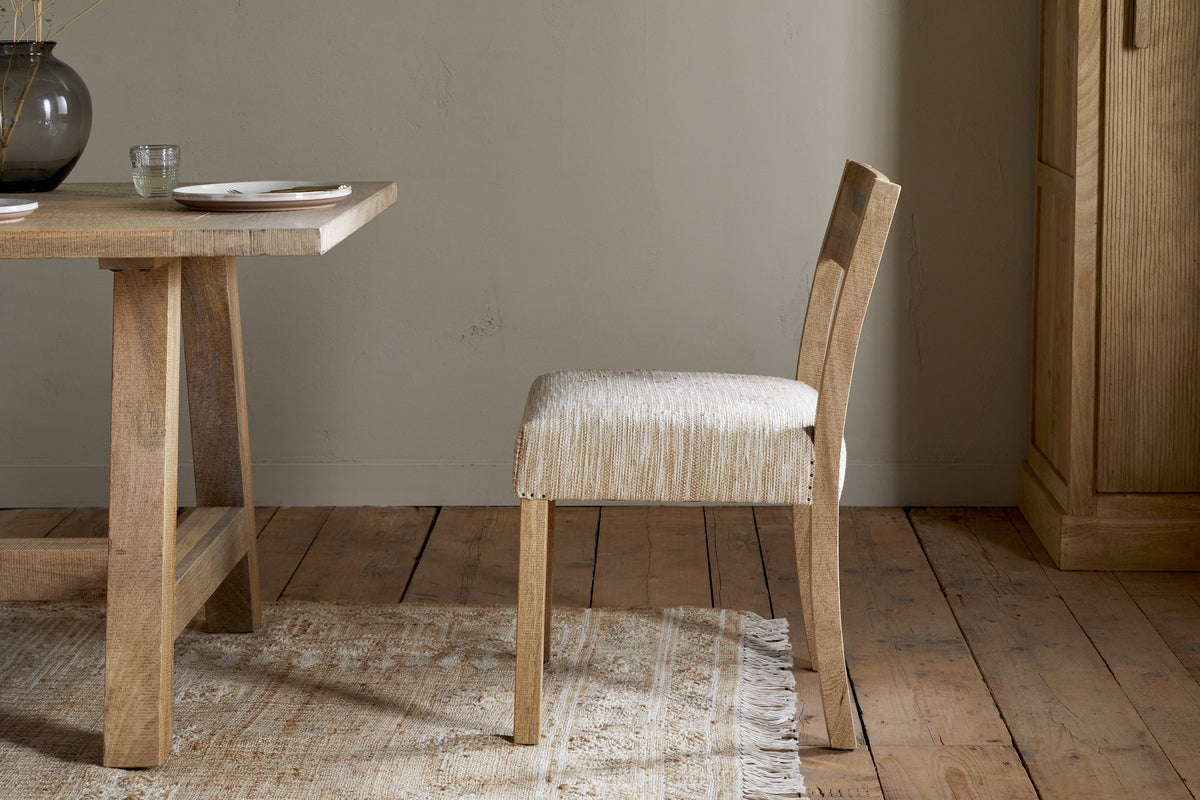 The Zoha Cotton & Jute Upholstered Dining Chair - Natural by nkuku features a textured beige seat and backrest beside a matching wooden dining table, set on a light rug over wooden floors, with a cupboard and ceramic vase in the background.