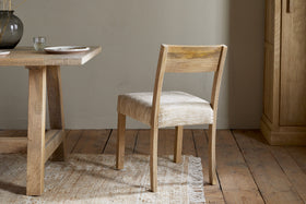 The Zoha Cotton & Jute Upholstered Dining Chair by nkuku, in a natural finish, sits on a textured rug next to a wooden dining table. Plates and a glass vase are on the table, with wood flooring, neutral walls, and a wooden cabinet in the background.