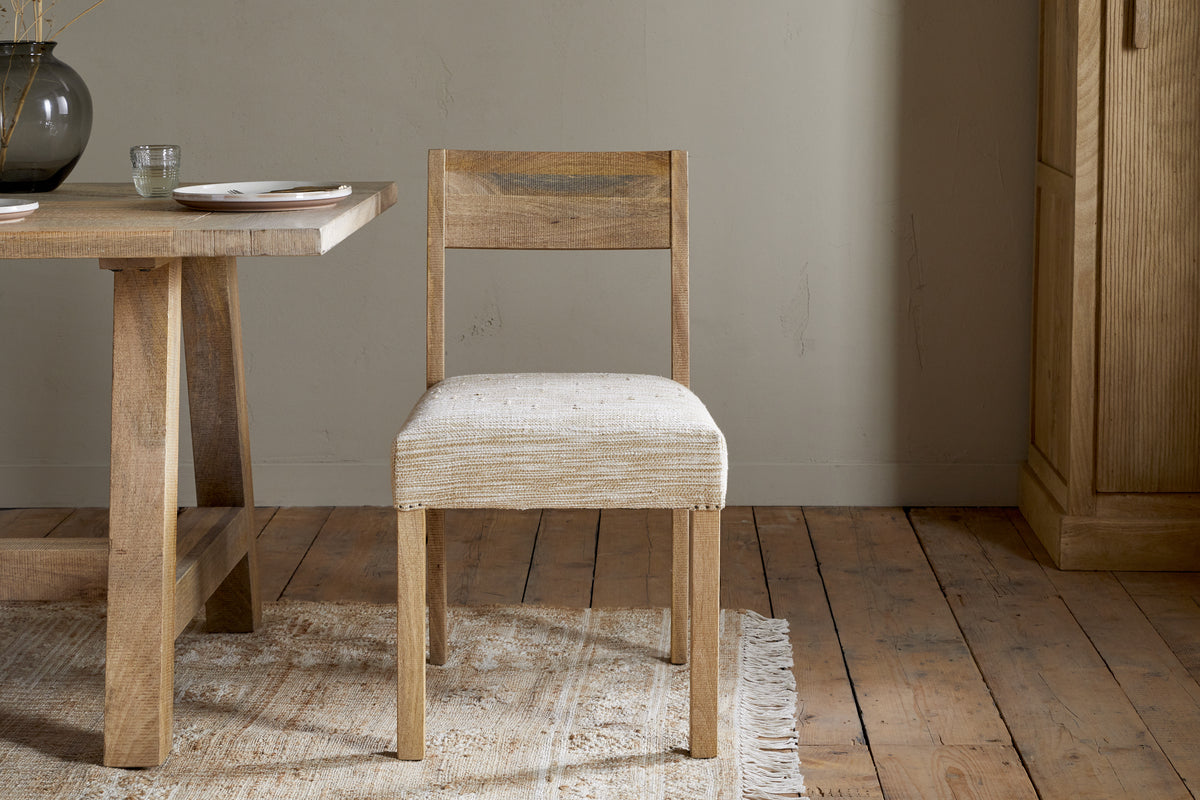 The Zoha Cotton & Jute Upholstered Dining Chair by nkuku sits on a textured rug beside a wooden dining table. The space features wooden floors, neutral walls, and minimalist decor with a glass vase and plates on the table.