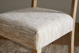 The nkuku Zoha Cotton & Jute Upholstered Dining Chair - Natural features a handcrafted mango wood frame and a textured, off-white cotton and jute seat, pairing elegantly with patterned rugs and wooden floors for an inviting dining space.